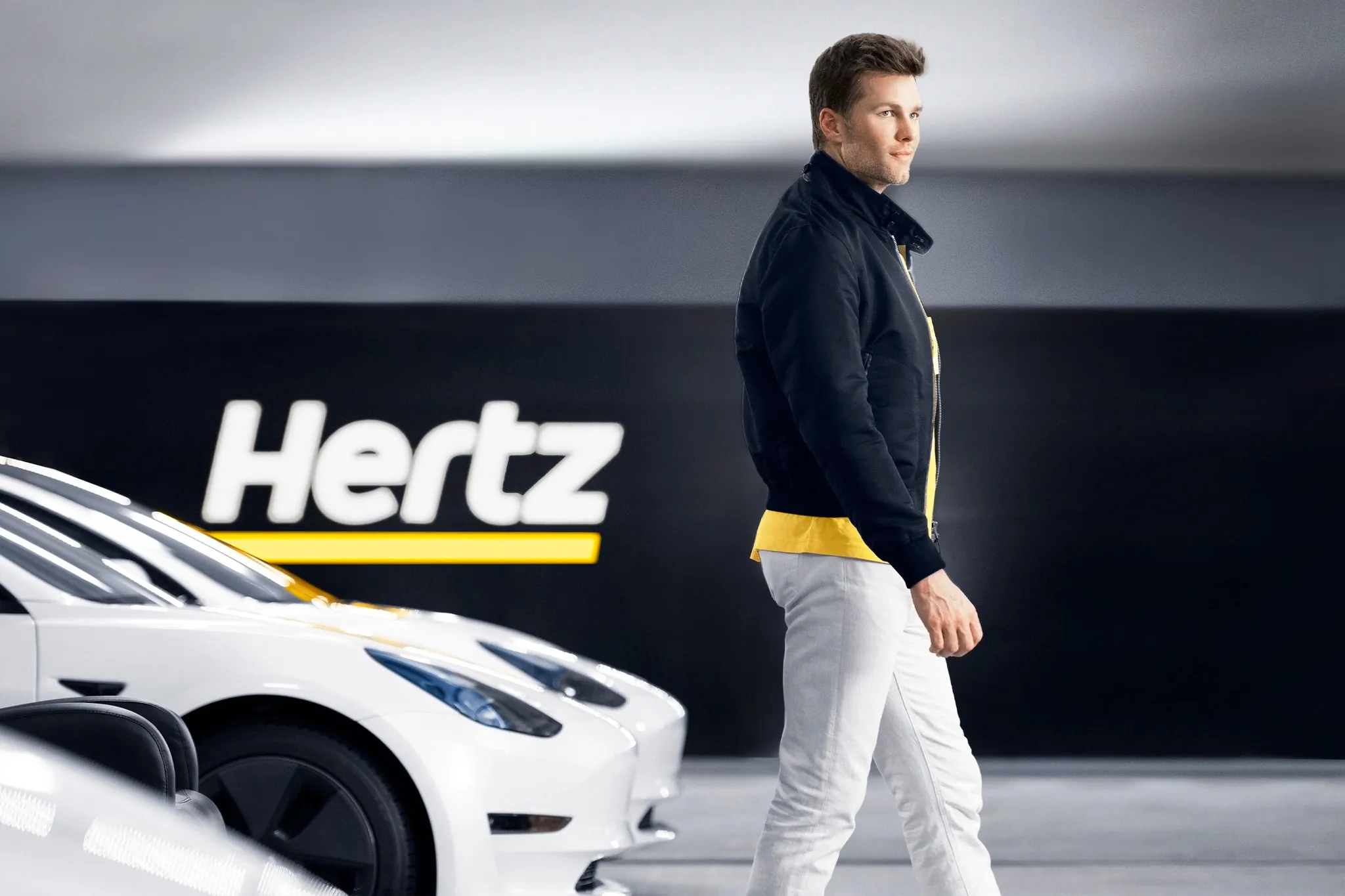 Tom Brady standing next to a Tesla vehicle in a Hertz rental lot